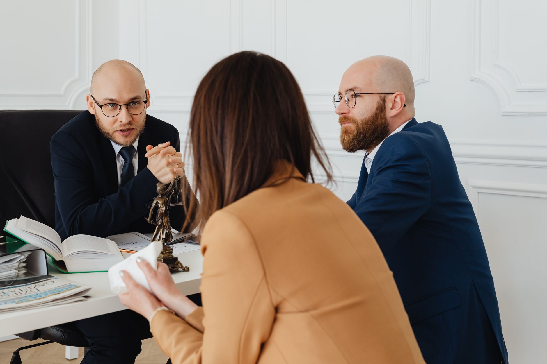 Two lawyers in suits meeting a client across a desk with legal books and a scales-of-justice statue